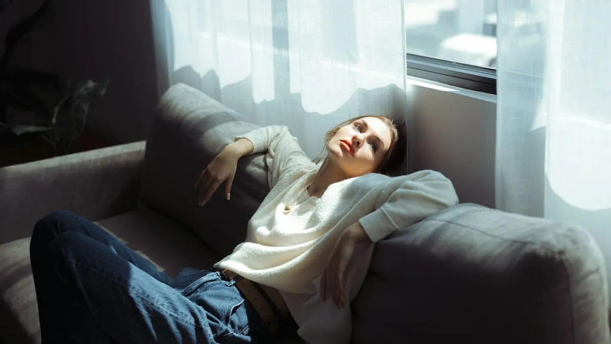 Опущение матки и стенок влагалища: упражнения и что делать. A woman in a white sweater and blue jeans reclining on a grey sofa Фото: James Forbes, Unsplash